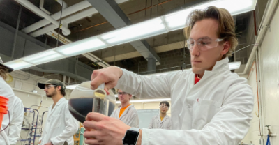 Student in a lab coat pouring a solution into a vessel.