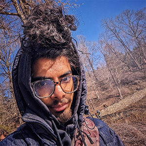 man with a glasses and a sweatshirt standing in the middle of a forest after fall. 