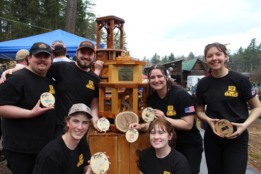 Group of six smiling young men and women in black T-shirts holding round medals standing by a large wooden trophy.