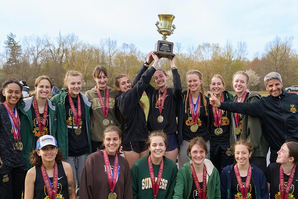 Fifteen young women and one man in two rows holding up trophy and wearing medals.
