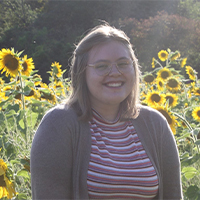 Maddie smiling. She is wearing a striped top and a sweater. She is standing in a sunflower field
