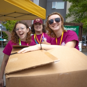 3 student helpers in maroon shirts holding a cardboard box at the camera