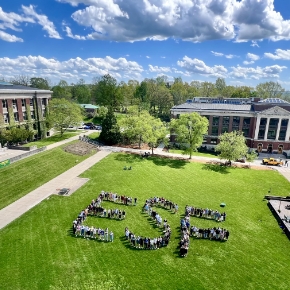 Aerial view of campus quad with green grass and blue cloud dotted sky, students lined up in letters E S F.
