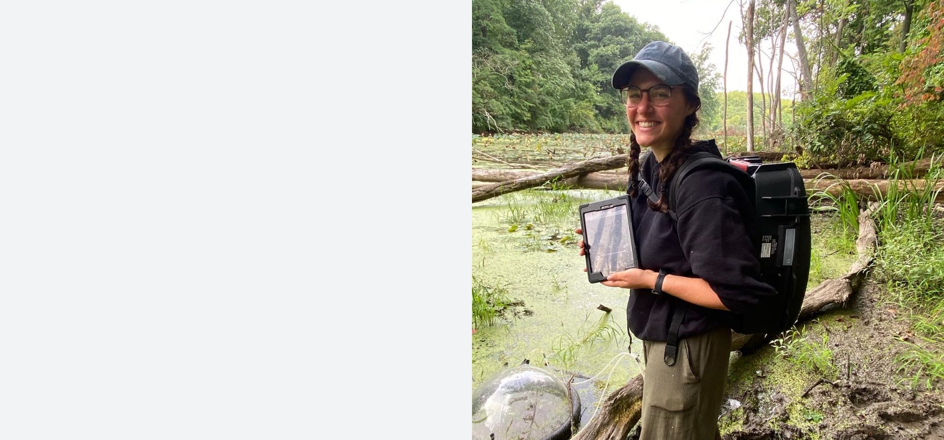 Student wearing a cap and backpack in the field holding a tablet.