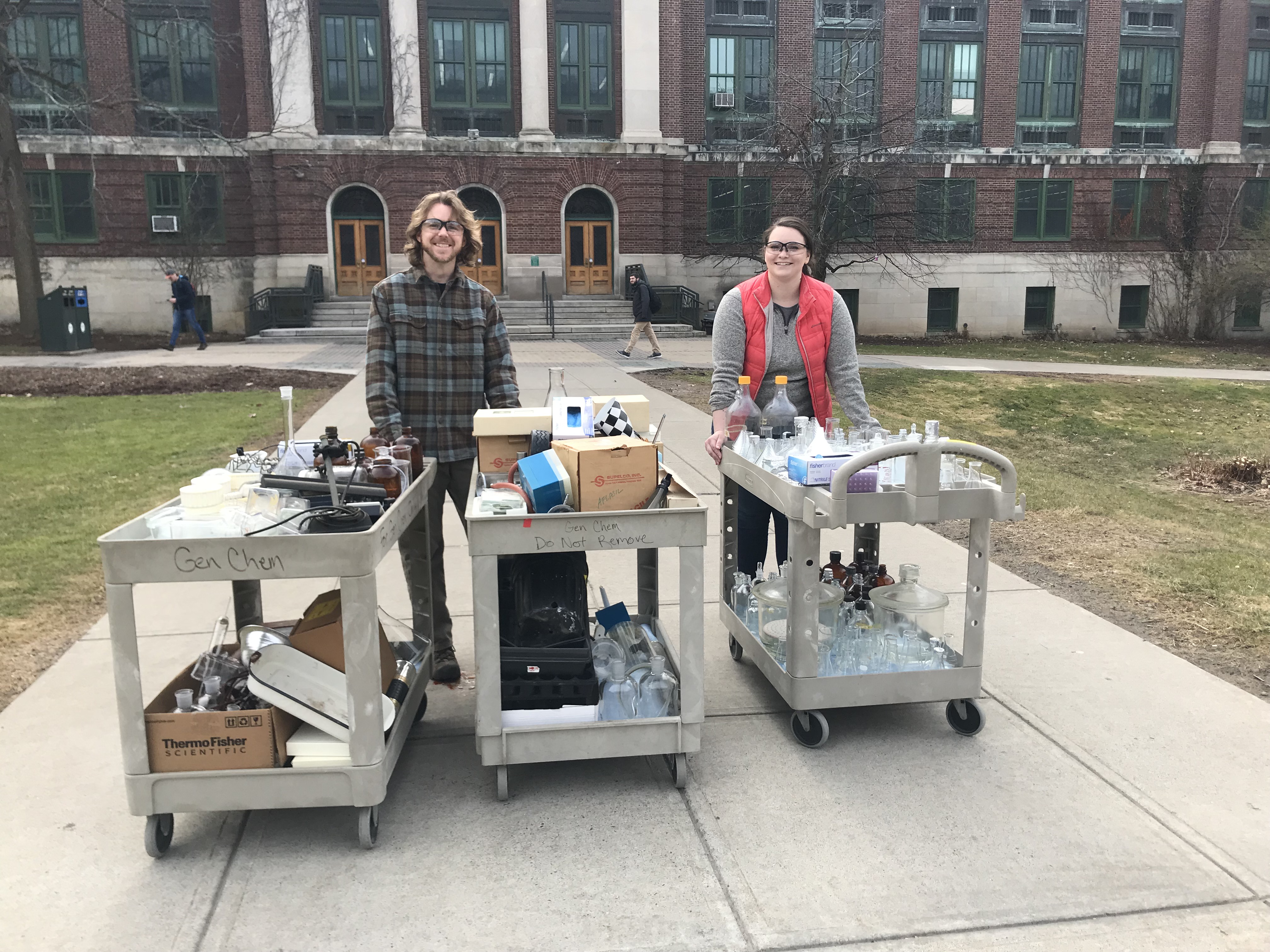 Two smiling people, transporting extra lab material to their lab on a cart