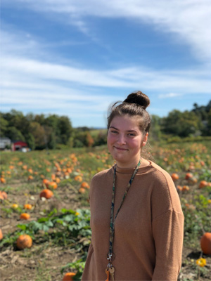 Jackie Whitehead in a pumpkin patch. She is wearing a brouwn sweater and her hair is in a bun.
