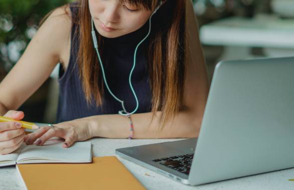 Student writing in a notebook at a desk next to a laptop computer.