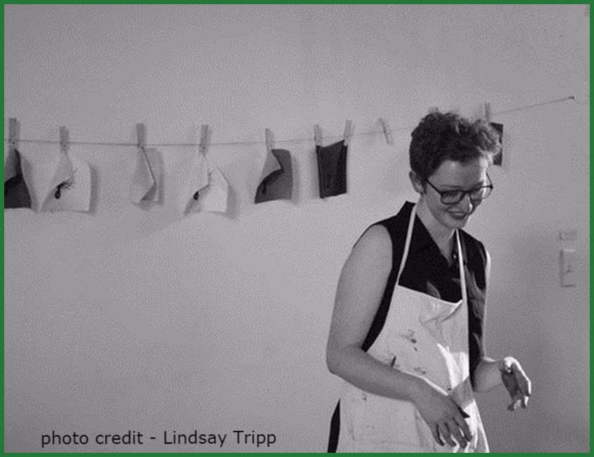 black and white image of Emma Percy. She is wearing an apron and there a small pieces of clothes hanging in a clothes line behind her. Photo credit by Lidsay Tripp