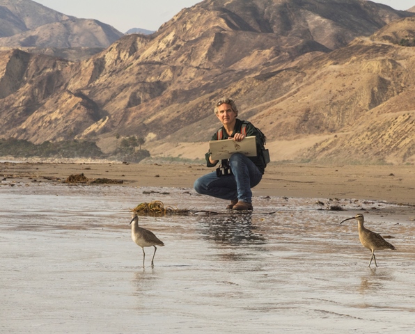 Niel Rizos kneeling on a river side with mountains on the back drop. He is painting.