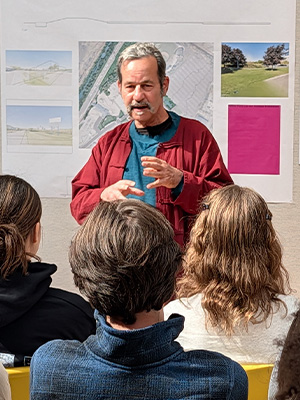 Community member wearing a red and blue shirt, sharing his perspective in a classroom. There are posters and maps behind the community member.