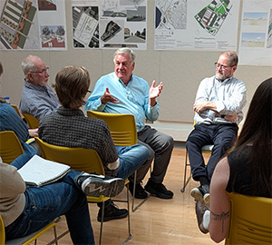 Members from local agencies and firms talking with each other.  They are sitting on yellow chairs in the spacious, dome-lit, white-board adorned Jury Crit room in Marshall Hall.