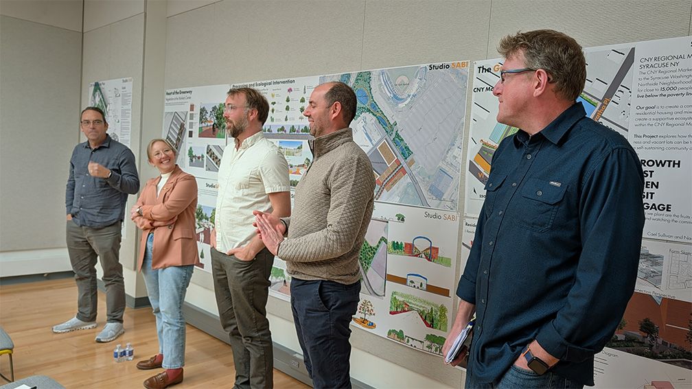  Five leaders from the Central New York community stand next to each other and address students. They are flanked by informative wall posters&nbsp;that explain landscape design and possibilities