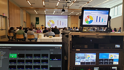 A chart for greenhouse gas emmission reduction requirements shown on a screen at Gateway and a monitor showing the same slide. Recording equiments at Gateway