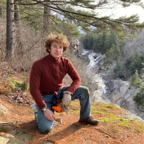 ian davis wearing maroon crew neck, jeans and hiking boots, kneeling on the ground. There is a creek behind him