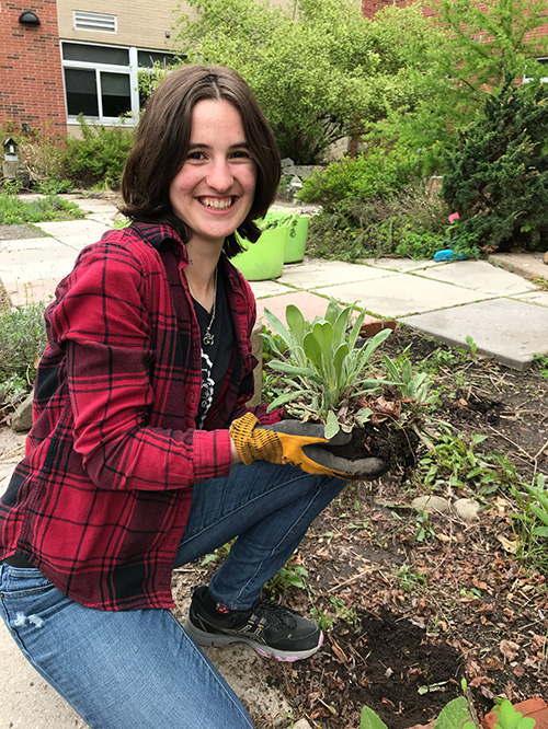 Josie outside planting a plant. She is wearing a red and black plaid shirt and jeans. She has gardening gloves on. 