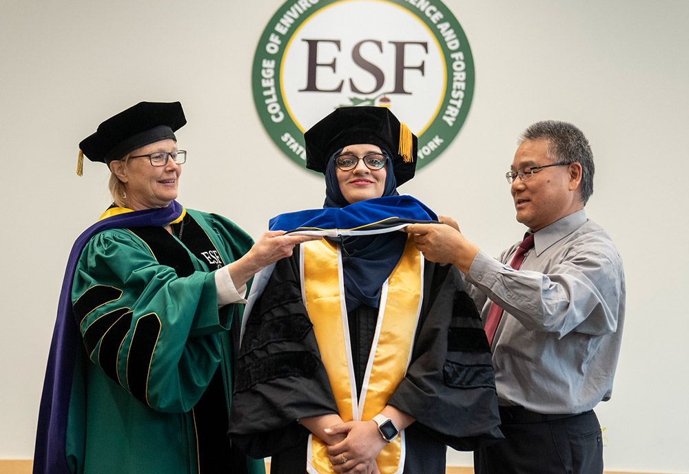 Female students being hooded by college president Joanie Mahoney (left) and male professor (right).