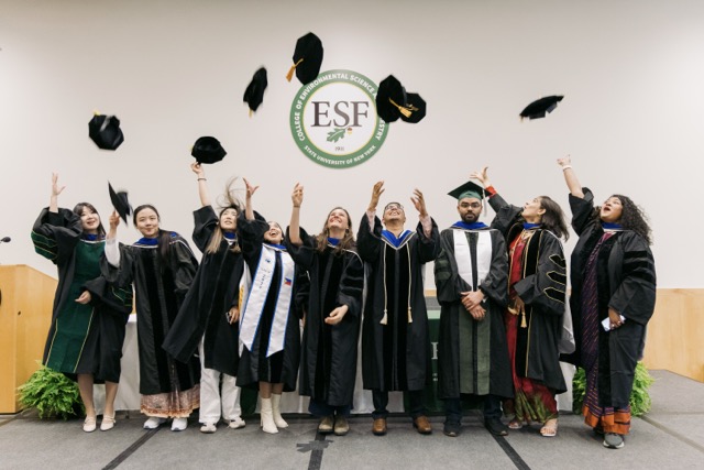 Group of just hooded higher degree recipients celebrating on stage throwing their caps in the air.