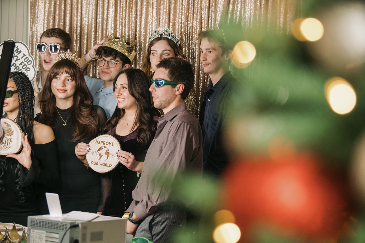 Group of students posing in a photobooth with props looking off screen to the left at a camera