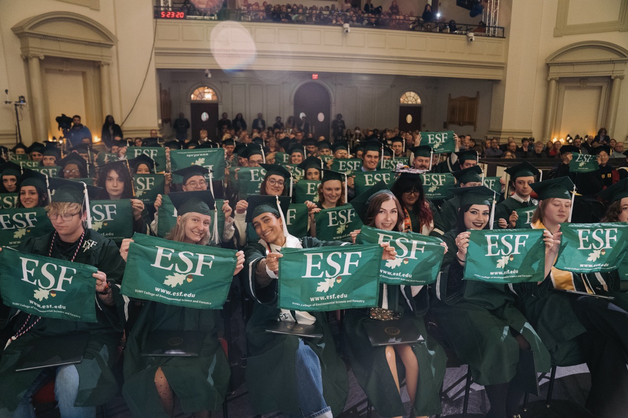 Students in regalia, seated, holding up ESF flags at Commencement