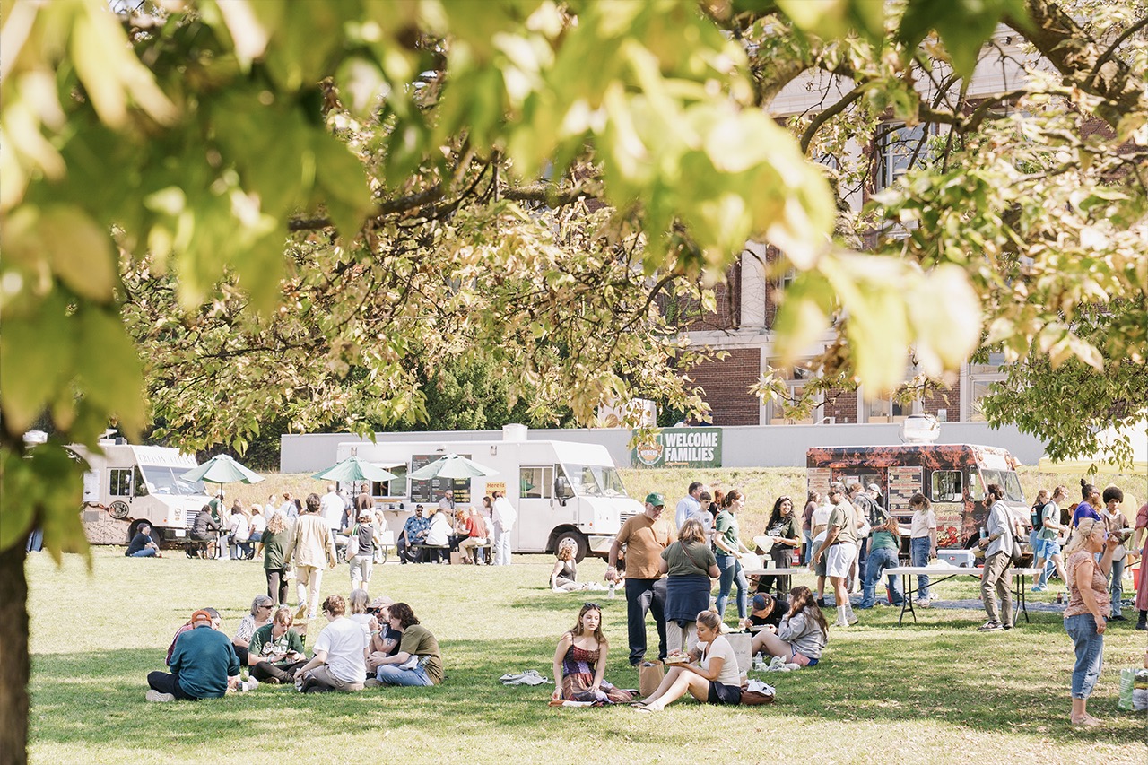 Photo of the quad through green tree leaves on a brigh sunny day with families scattered about. Bray Hall in the background, food trucks line the grass edge.