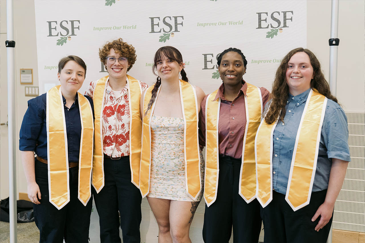 Group of five students posed together in a line looking at the camera in front of step and repeat esf logo banner
