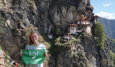 Gillian Hall stands holding an ESF flag outside Tiger's nest Monastery in Bhutan.