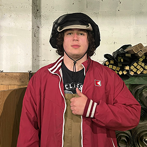 Man in a hat giving the thumbs up. There is lumber in the background. 