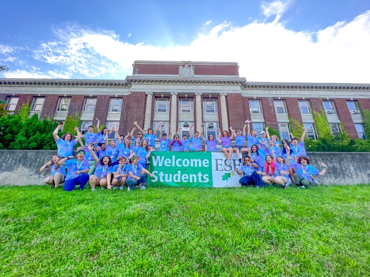 Group of students energetically waving at the camera around a banner that says Welcome Students with the ESF logo. There is a building in the background that is brick with pillars and has a bunch of windows. 