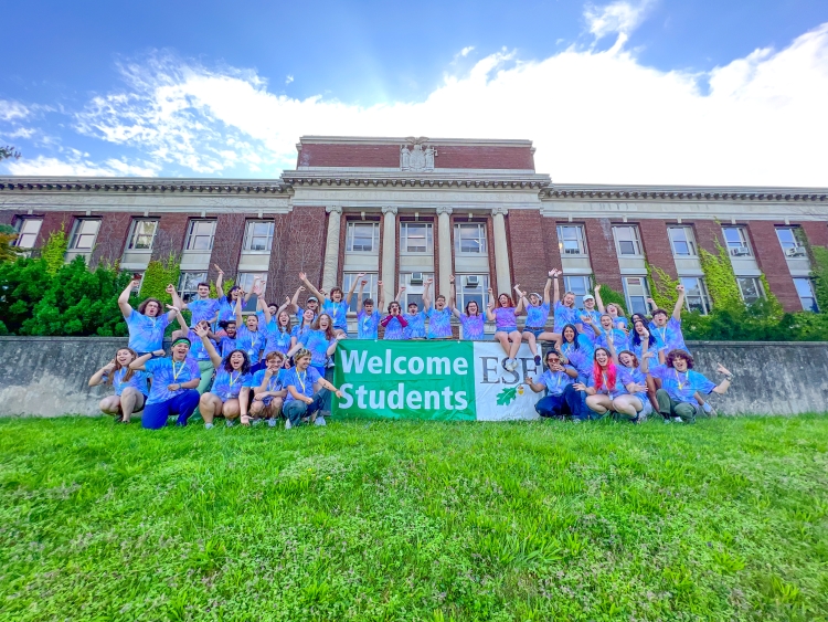 Group of students energetically waving at the camera around a banner that says Welcome Students with the ESF logo. There is a building in the background that is brick with pillars and has a bunch of windows.