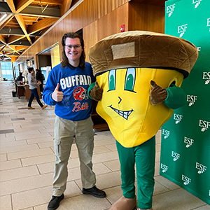 Man with glasses and a buffalo bills shirt on standing next to Oakie the Acorn. 