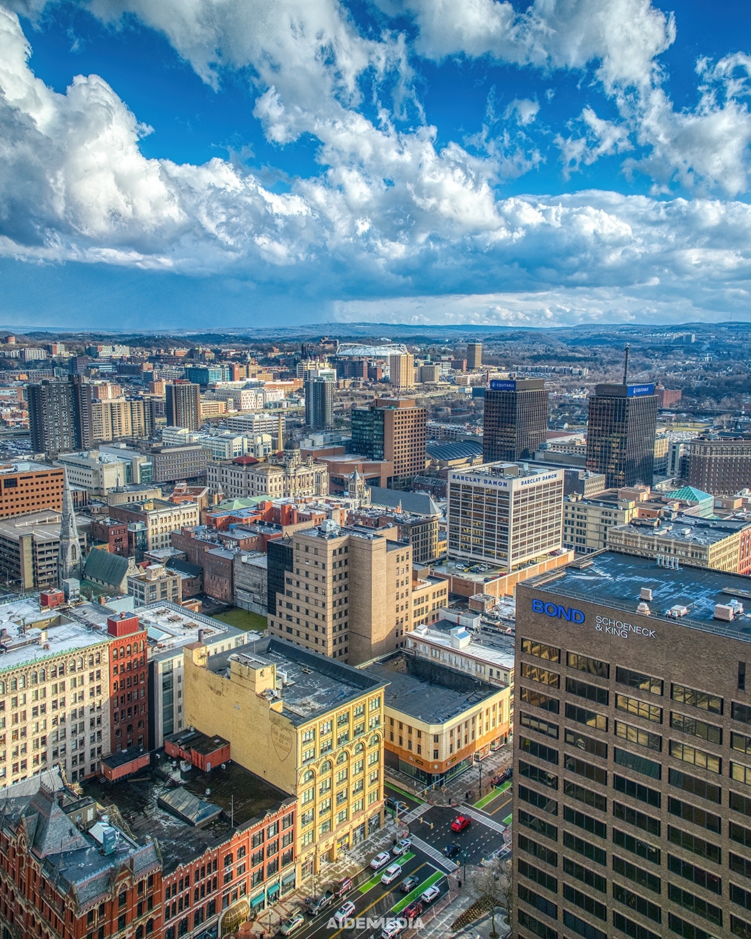 A city of a blue skied day. There are many buildings of various heights and sizes smattered throughout the picture. 