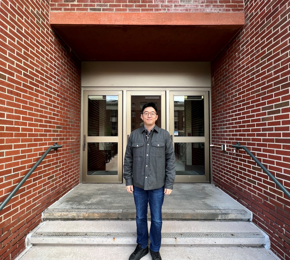 Man wearing grey shirt and blue pants standing on steps in front of brick doorway