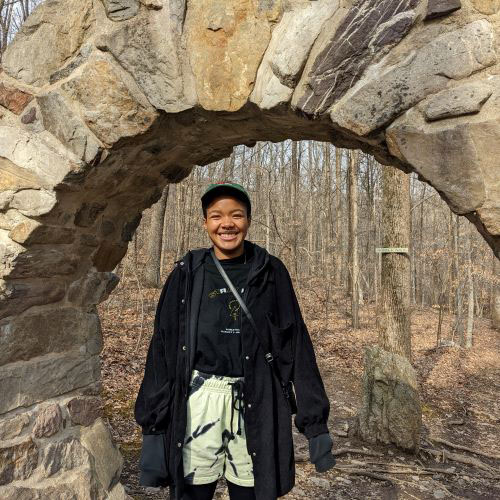 Woman standing in a stone arch with a bare leaf forest in the background. 