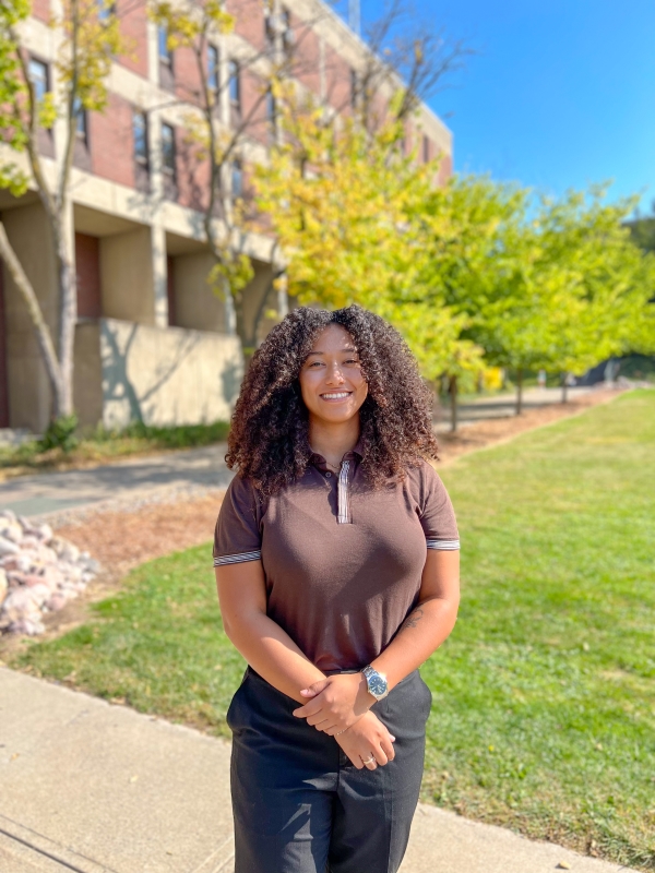 Woman with long black curly hair and a brown shirt standing in front of a green grass space with trees in the background. The trees have leaves and you can see people walking in the distance. 