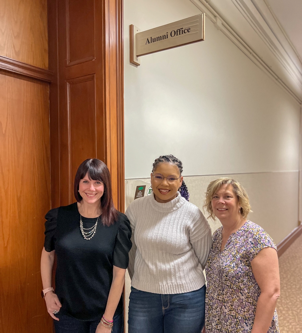 Three women standing in front of a doorway with a sign that says Alumni Office above them. 