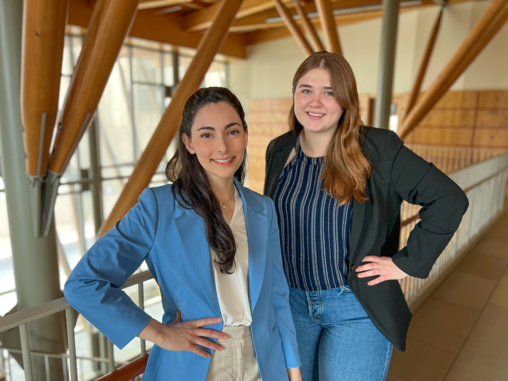 Two women standing on a concourse that has lots of brown panneling and railings in the background. They are both wearing blazers that range from light blue to dark blue. 