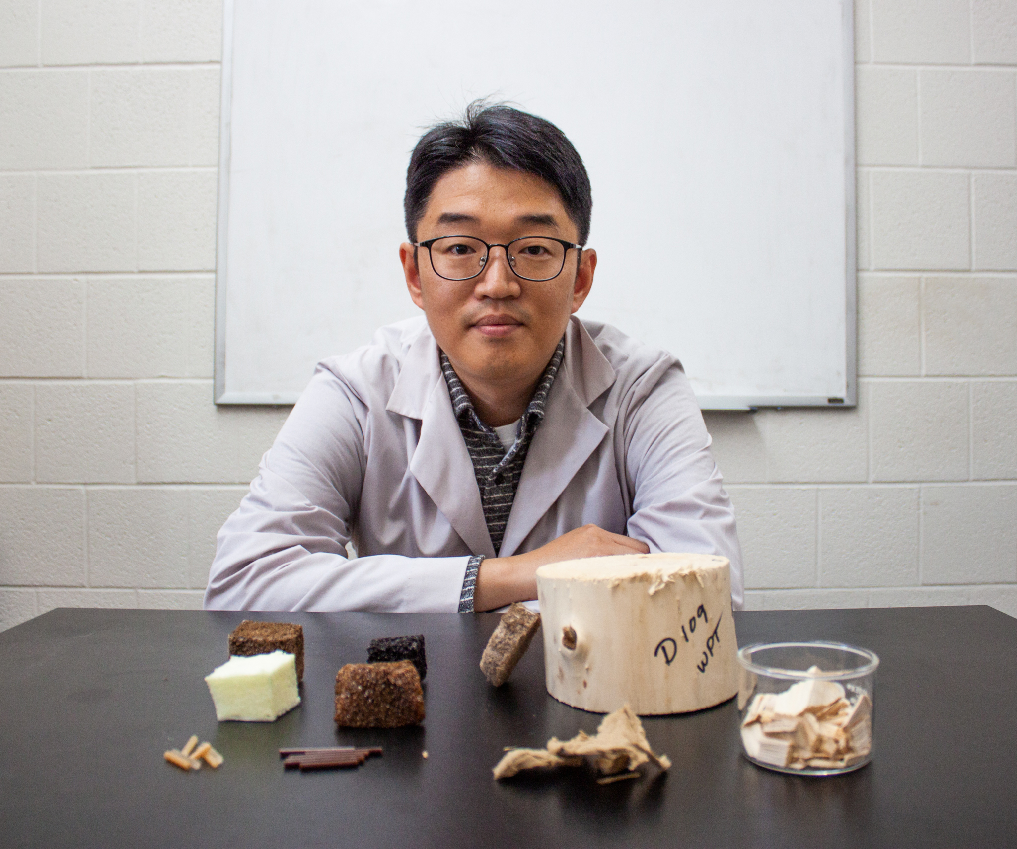 Man with a lab coat sitting with various wooden and foam materials in front of him.