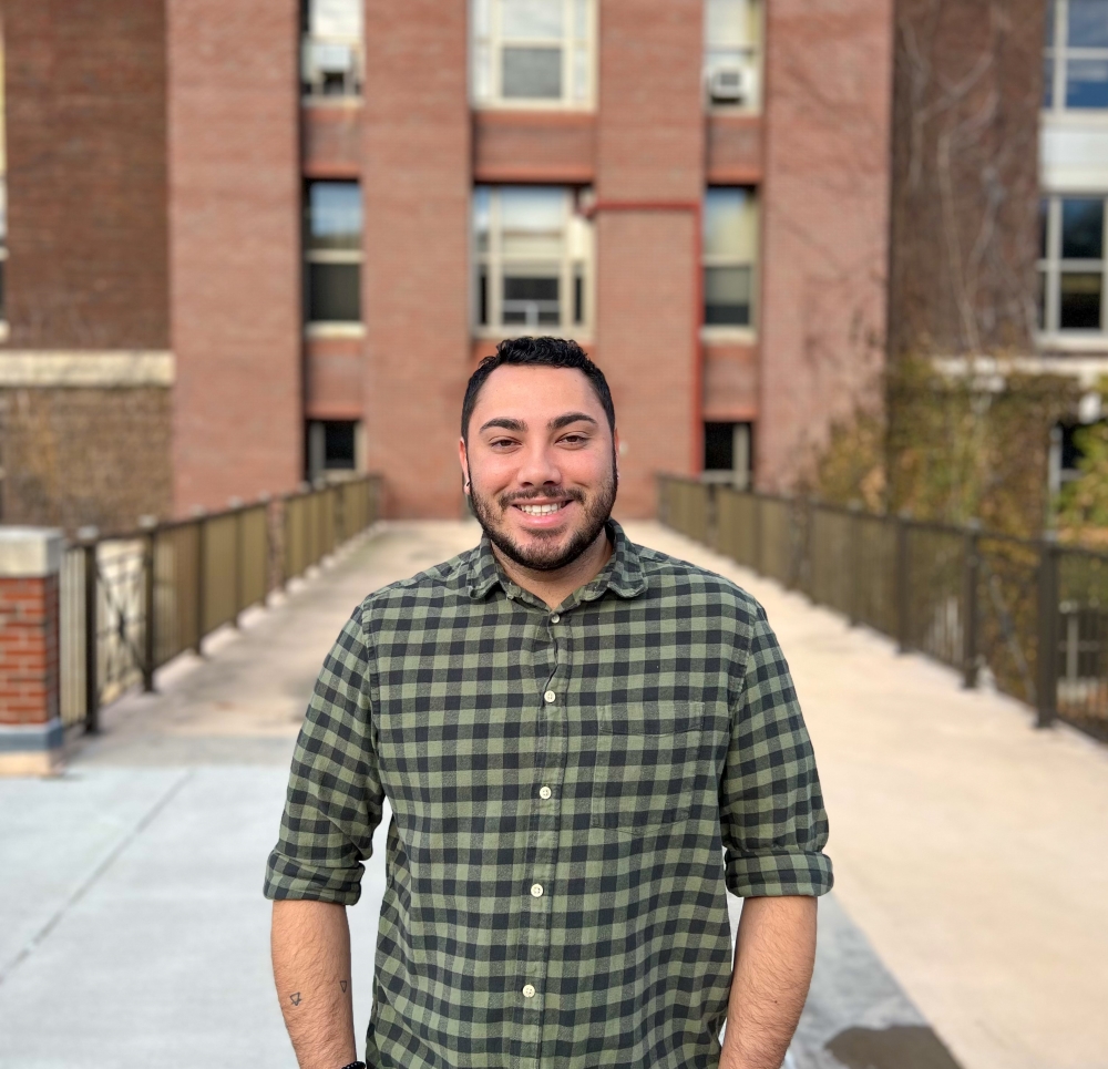 Man in a plaid flannel shirt standing in front of a brick building with green ivy on the sides. 