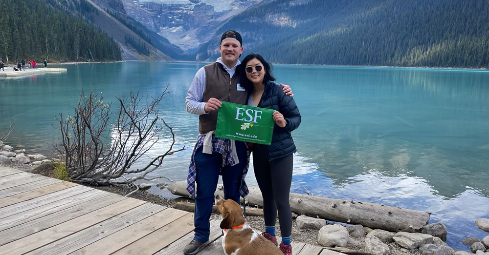 Man and woman standing in front of a lake surrounded by mountains. They are holding an ESF flag. 