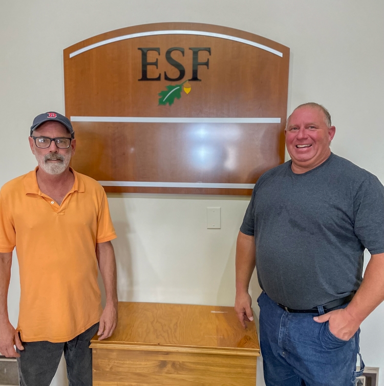 Two men standing in front of a wooden sign. 