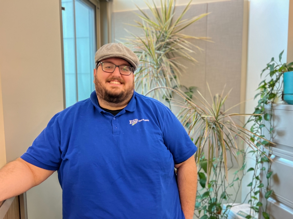 Man in a blue shirt, glasses and a hat standing in front of a bunch of plants.