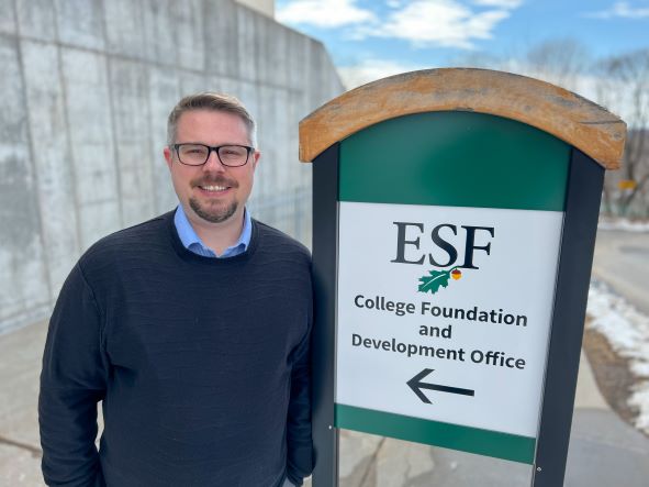 Man with a sweater and a colored shirt standing next to a green sign that has the ESF logo and the words: College Foundation and Development Office with an arrow pointing to the left. 