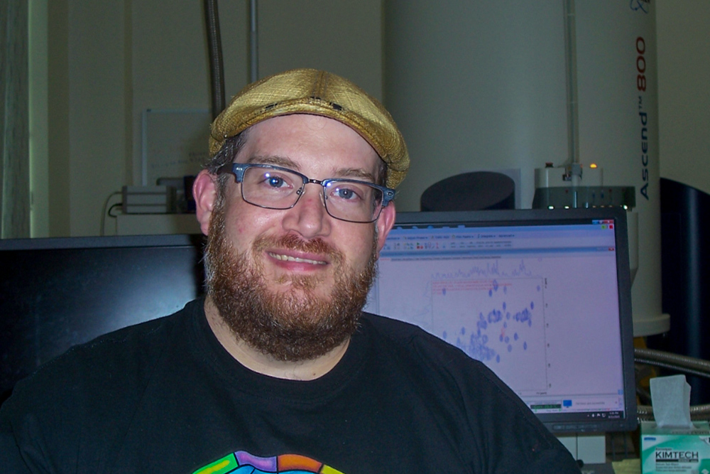 Man with beard and mustache wearing a hat sitting in front of a computer screen. 