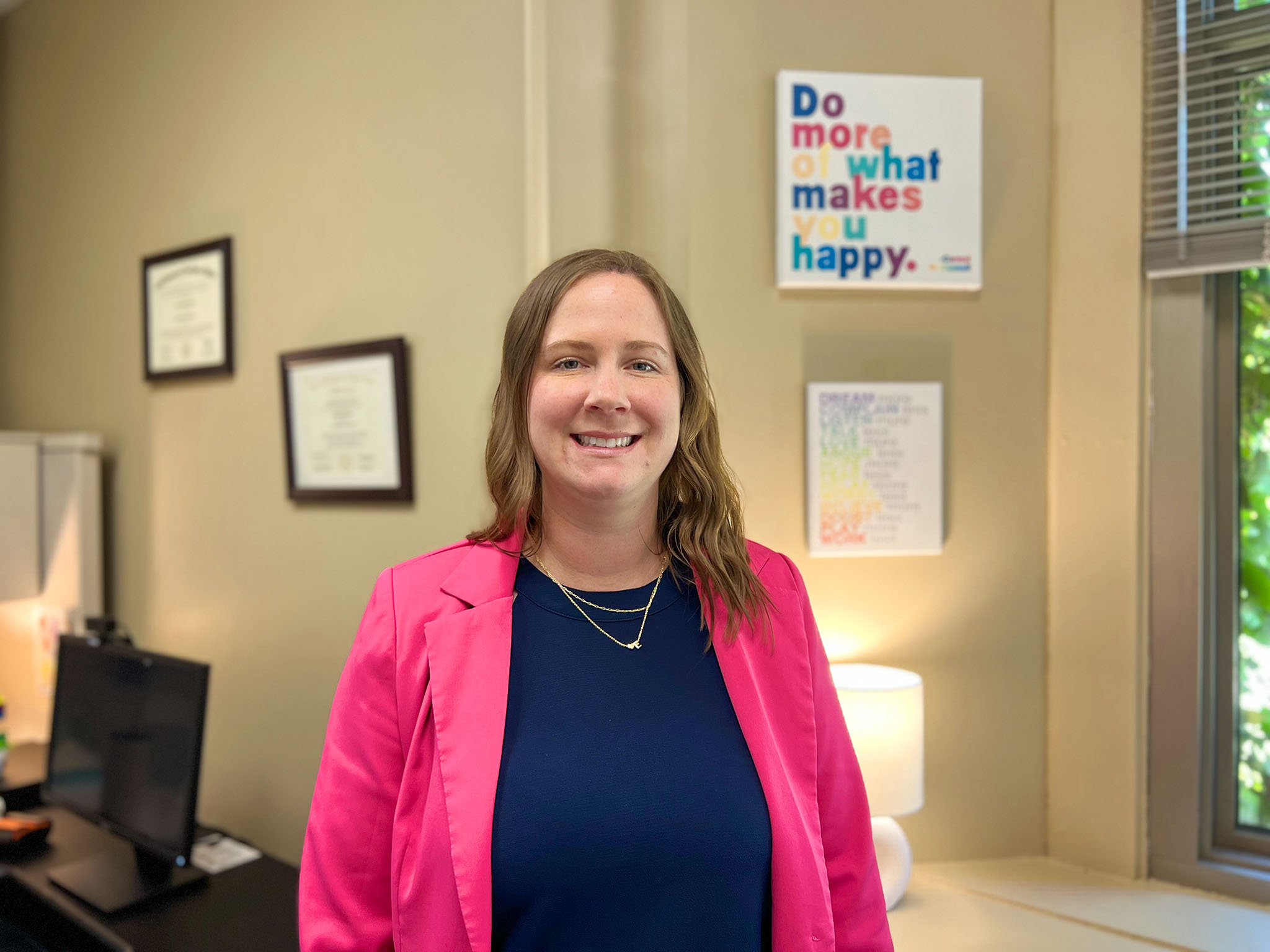 Woman in blue shirt and pink jacket standing in an office with a sign in background that says Do more of what makes you happy.