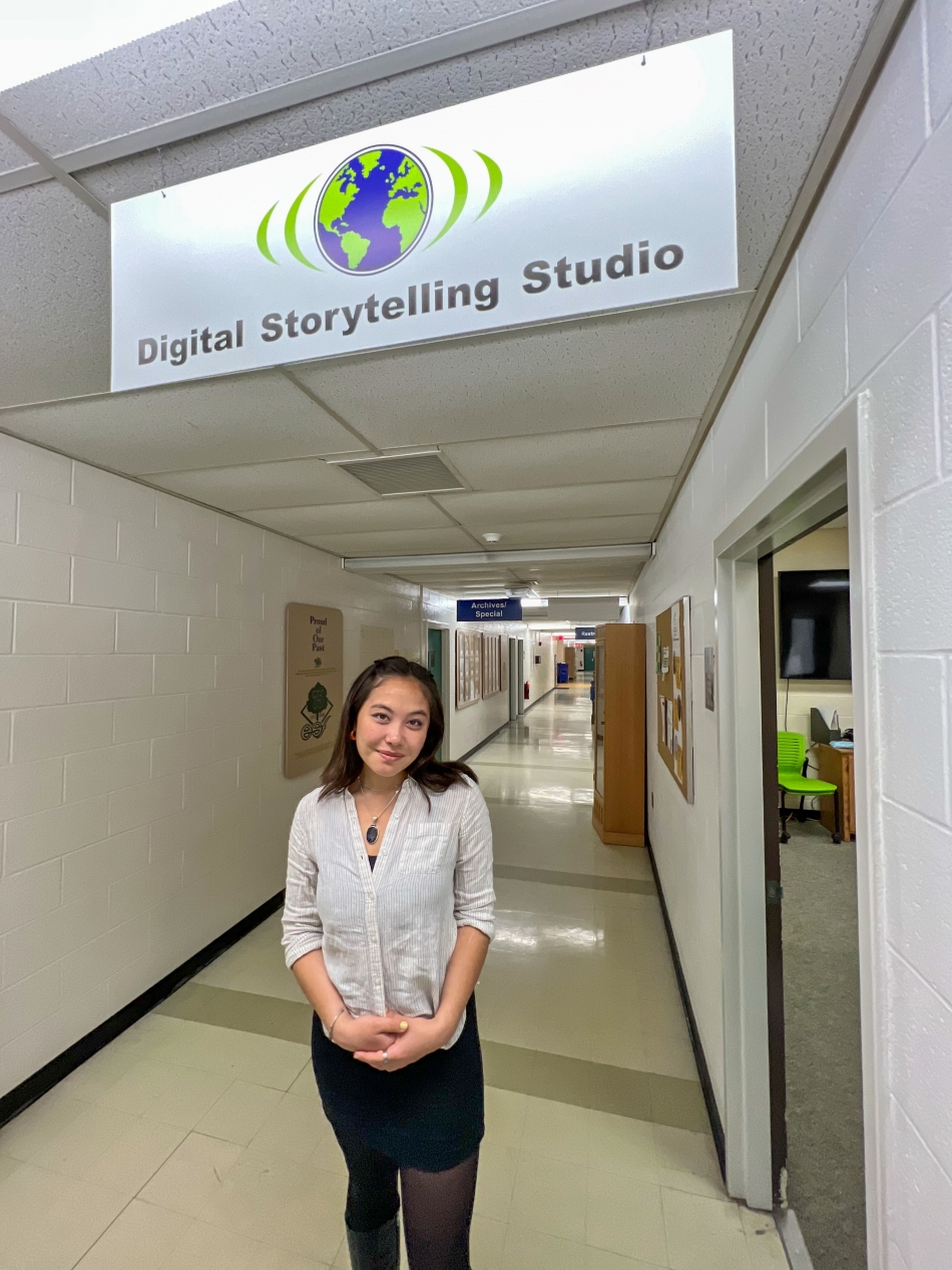 Woman standing in a hallway that's mostly white. There's a sign above her with a transmitting green and blue earth that says Digital Storytelling Studio below it.