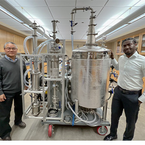 Two men standing next to a silver machine with multiple tubes and vats. 