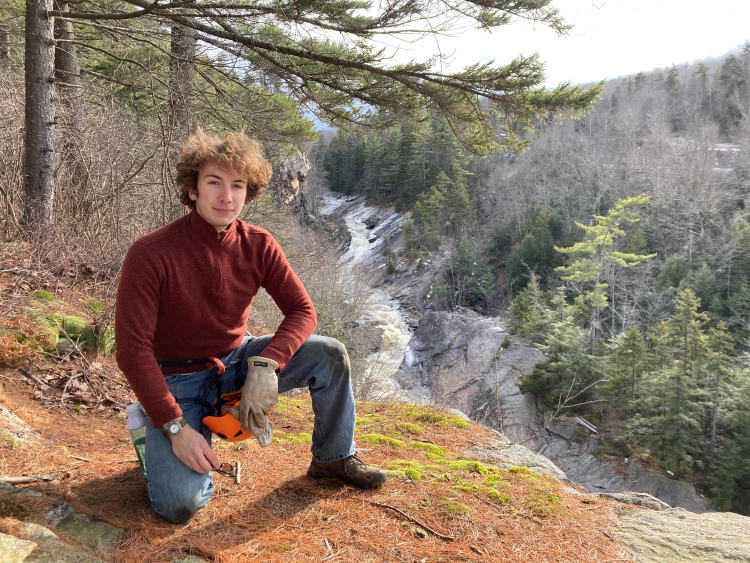 Man crouching down on the ground. He's in nature with a long gorge like stream in the background. Many of the leaves have fallen off the trees. 