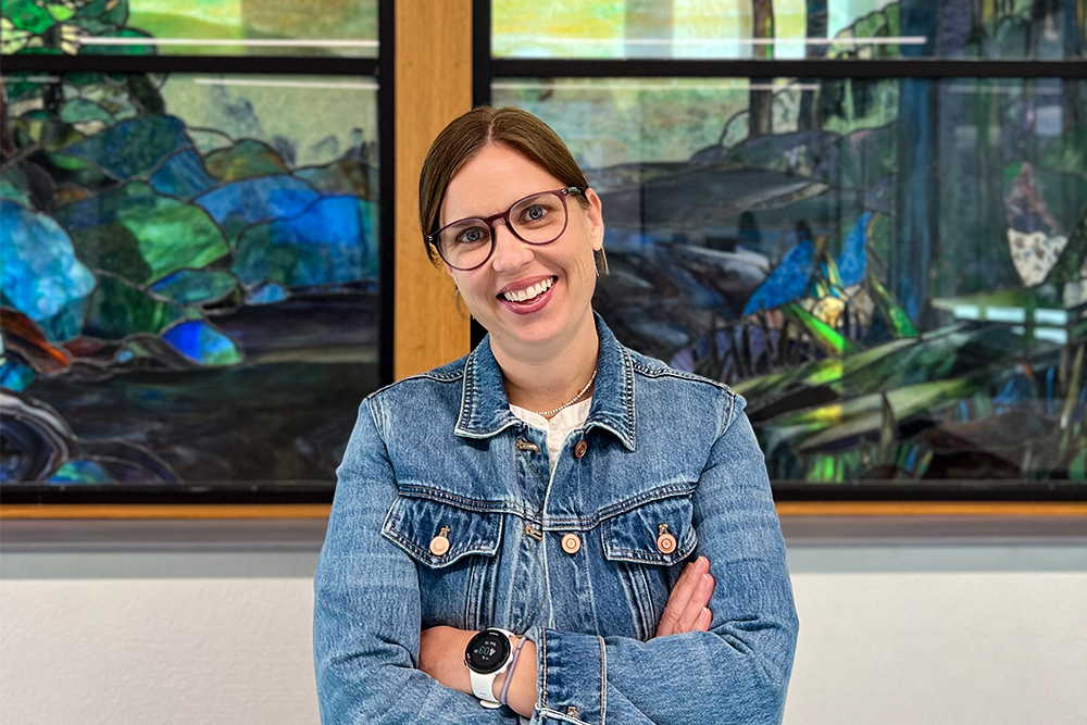 Woman wearing denim jacket standing with arms crossed in front of a stained glass window.