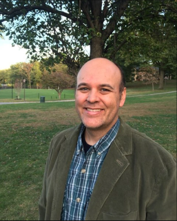 Man wearing blue plaid shirt and olive jacket standing in front of tree