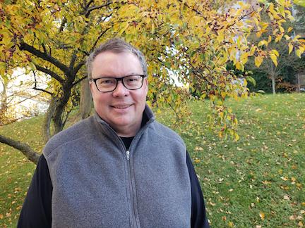 Man wearing glasses a long-sleeved black shirt and grey fleece vest standing in front of a tree with yellow autumnal leaves.